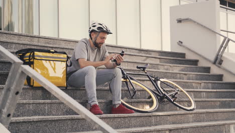 un repartidor de comida feliz sentado en un escalón junto a una mochila térmica responde una llamada telefónica