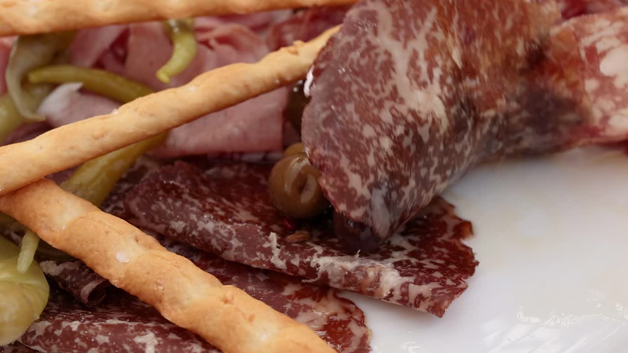 A fork picks up salami and olives from a plate with breadsticks and pickles.