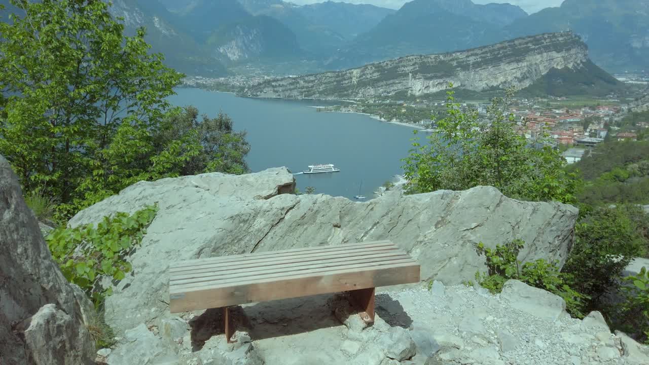 Vista em paralaxe de céus tempestuosos sobre o Lago de Garda, de Riva del Garda a Nago-Torbole, com bancos panorâmicos e vegetação exuberante.