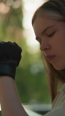 joven adulto con guantes negros rasca el codo suavemente, la luz del sol se filtra a través de hojas exuberantes creando un brillo cálido, el fondo borroso presenta árboles y vegetación