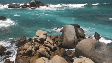 Blue-sea,-waves-crashing-against-a-stack-of-rocks-on-a-cliff-in-Tayrona-Park,-Colombia