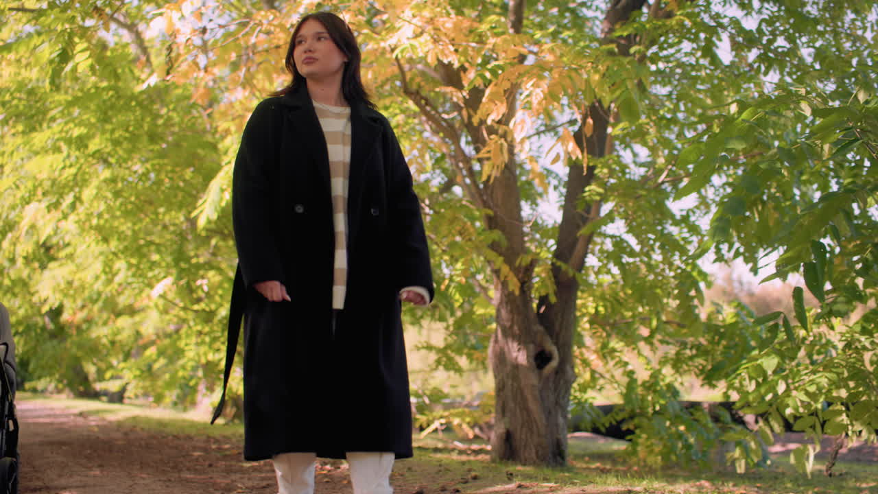 Una giovane donna riflessiva in cappotto invernale, top a righe, pantaloni bianchi e stivali neri cammina lungo il sentiero di un arboreto, ammirando il fogliame invernale sotto la luce del sole e l'ombra, con alberi che fiancheggiano i lati, in un'atmosfera di calma
