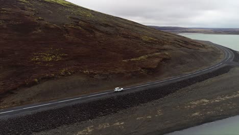 Toma-Aérea-De-Un-Automóvil-Conduciendo-Entre-Una-Montaña-Y-Un-Lago