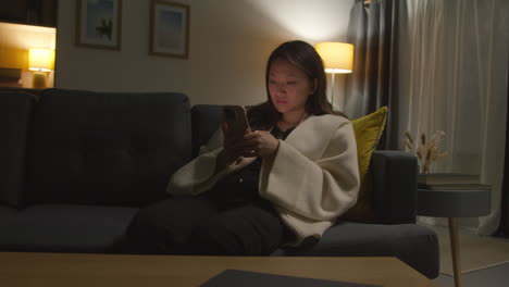 woman spending evening at home sitting on sofa with mobile phone scrolling through internet or social media 6