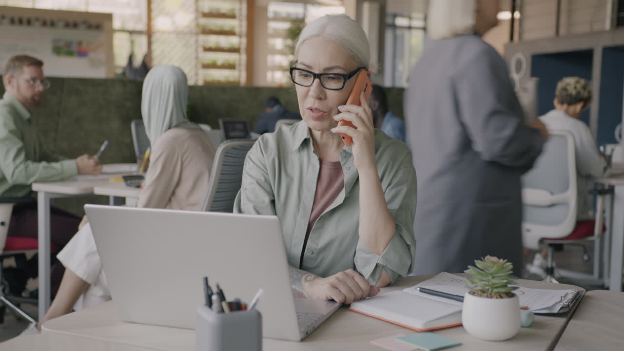 Senior Businesswoman Working on Laptop During Phone Call in Modern Office