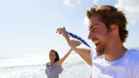 Vista-Lateral-De-Una-Joven-Pareja-Caucásica-Sosteniendo-La-Bandera-Estadounidense-Ondeando-En-La-Playa-4k