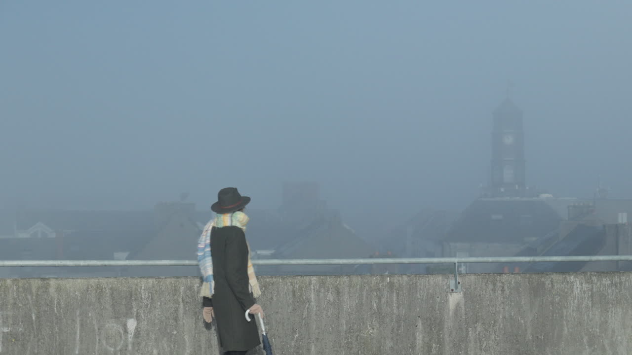 여자는 안개 낀 화창한 날에 도시의 감시탑을 바라보며 옥상을 가로질러 걷는다