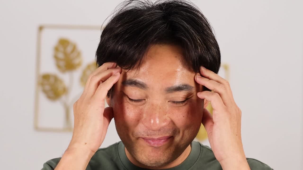 緑のシャツを着た中年アジア人男性が目を閉じてこめかみをこする。
