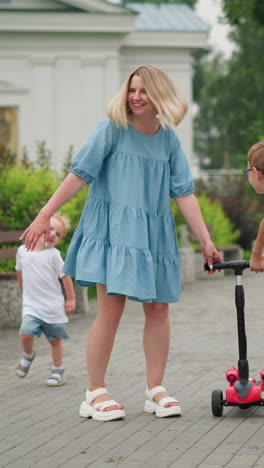 a mother walks alongside her son, who holds onto a scooter, in the background, the younger child runs happily to catch up to them as he hold his mum hand