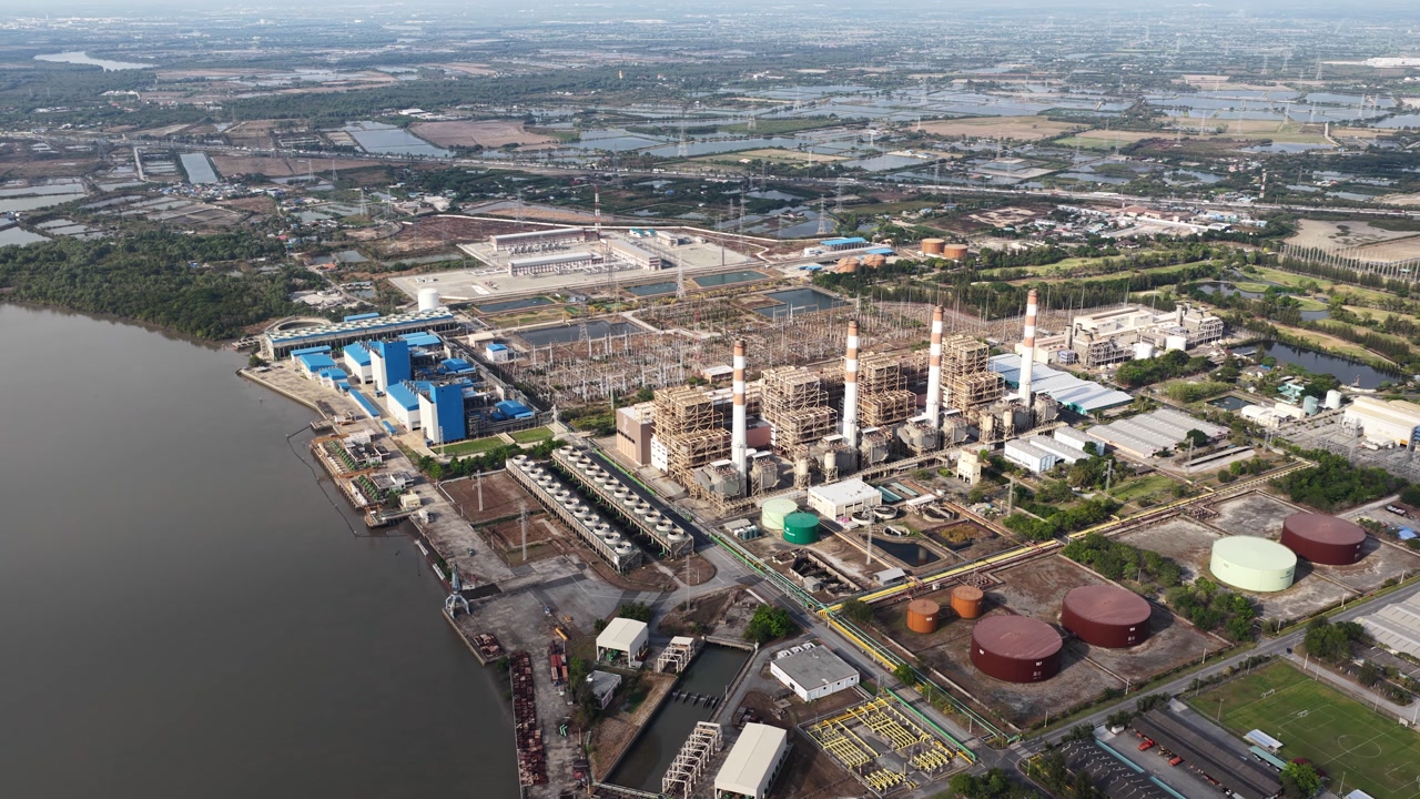 Expansive industrial zone featuring a large power plant with chimneys and cooling towers situated next to a major river, highlighting energy production and infrastructure in a developing landscape