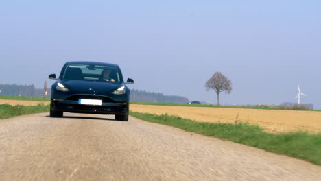 Tesla-Fährt-Auf-Einer-Kleinen-Asphaltstraße-Mit-Windrädern-Im-Hintergrund-Auf-Die-Kamera-Zu