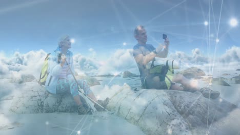 una pareja de ancianos caucásicos haciendo senderismo, tomando fotos con su teléfono inteligente, sobre las nubes y la red de conexiones.