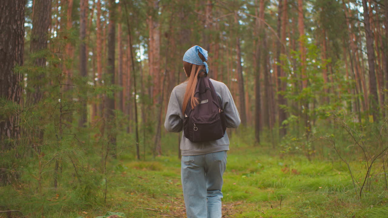 vista de la espalda de una mujer joven ajustando las correas de la mochila mientras camina a través de un camino pacífico y exuberante del bosque, con un bandana azul y rodeada de vegetación vibrante y árboles altos