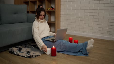 Young-woman-working-on-modern-laptop-while-sitting-in-a-cozy-room-around-candles
