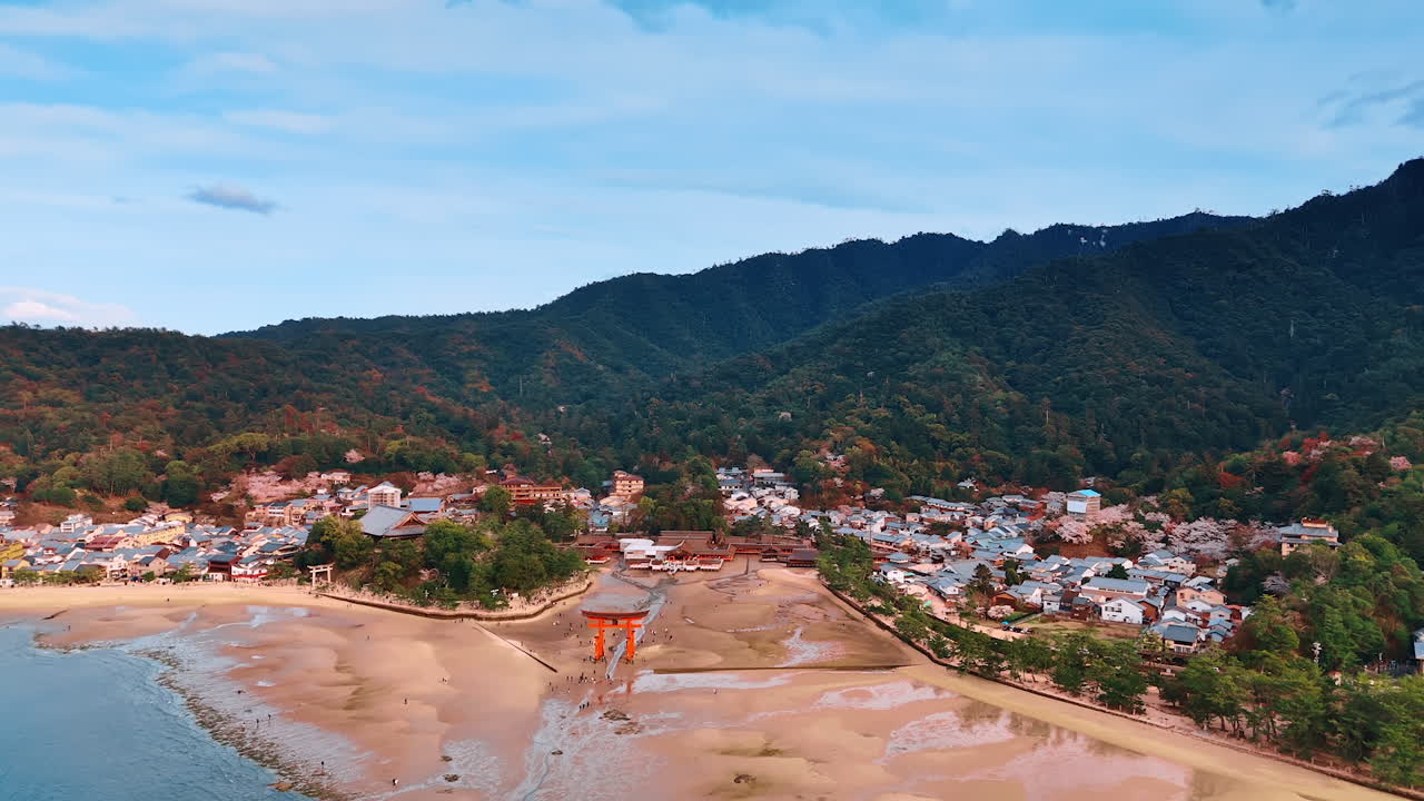 Sacred Itsukushima Shrine, Miyajima, Japan surrounded by private houses and green mountains. Tourists walk around red gates at low tide time. Aerial perspective.