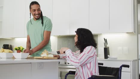 Happy-biracial-woman-in-wheelchair-using-tablet,-preparing-food-with-smiling-male-partner-in-kitchen