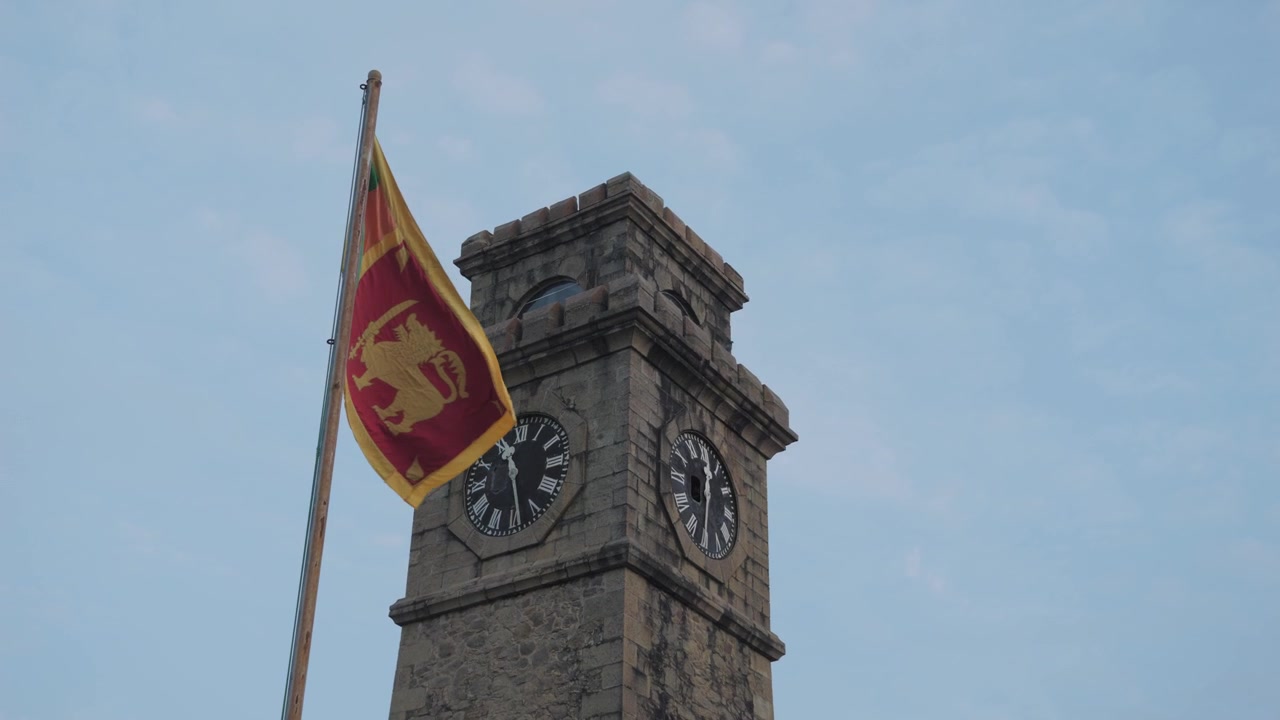 Low angle slow motion view of the Sri Lankan flag waving beside Galle Fort Clock Tower, showing historic stone architecture and clock faces against a pale blue sky.