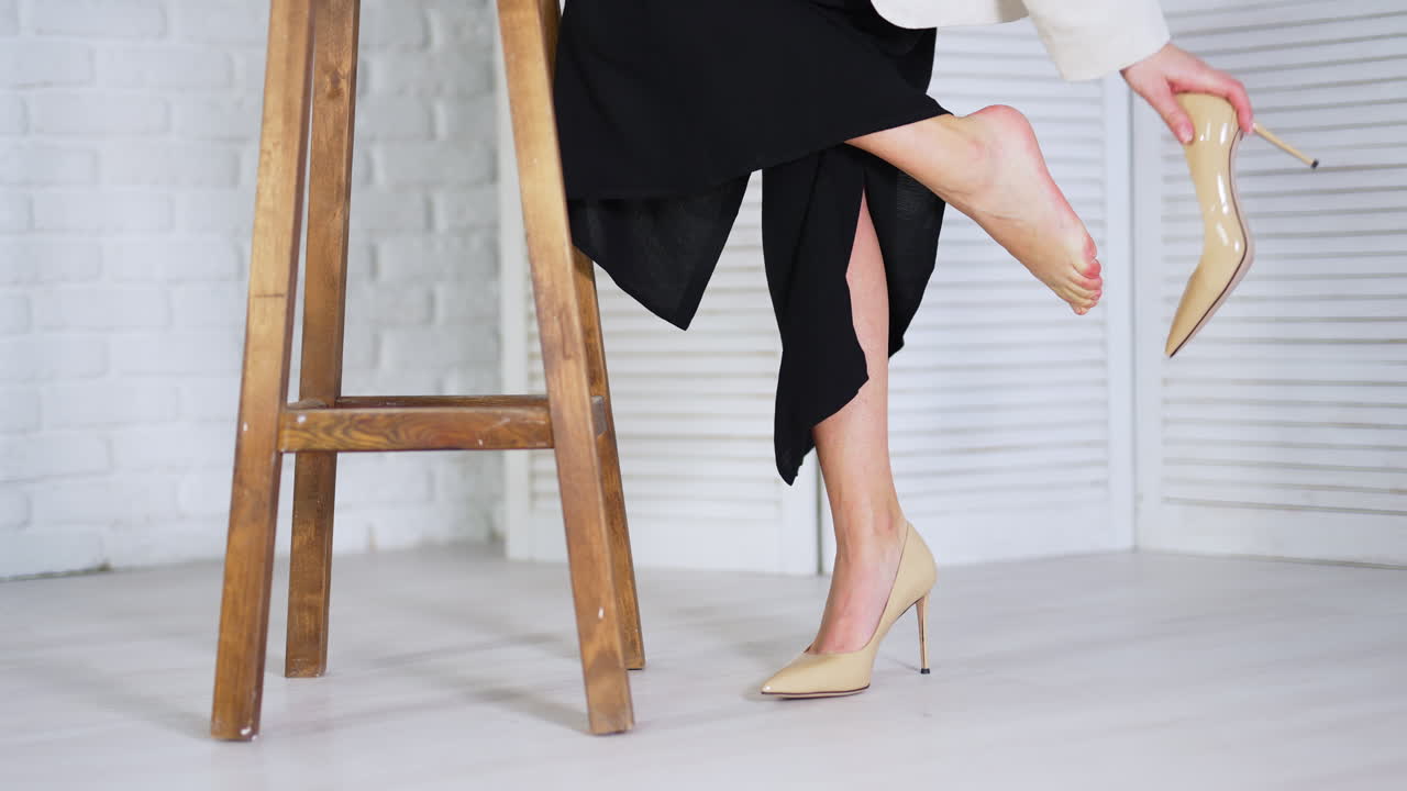 Lady takes off beige high-heeled shoe and feels relief. Lady massages tired foot leaning on a wooden tabouret. White backdrop.