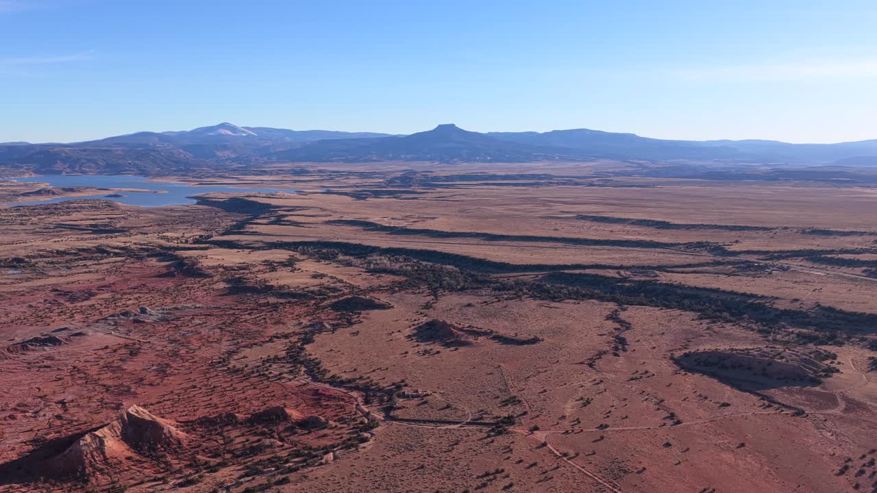 Drone footage moves steadily across expansive desert plains toward Cerro Pedernal, revealing winding land patterns, sparse vegetation, distant water, and layered terrain beneath clear blue skies