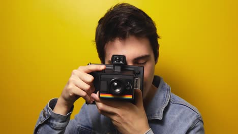 Portrait-of-attractive-young-millenial-man-taking-pictures-on-vintage-film-camera-on-yellow-background