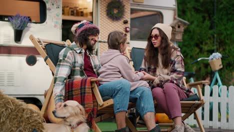 Un-Chico-Moreno-Feliz-Con-Una-Camisa-A-Cuadros-Verde-Acaricia-A-Su-Perro-Color-Crema-Y-Su-Novia-Se-Comunica-Con-Su-Pequeña-Hija-Que-Está-Sentada-En-El-Regazo-De-Un-Hombre-Cerca-De-Un-Remolque-En-El-Campamento-Durante-Sus-Vacaciones-Conjuntas-Y-Un-Picnic-Fuera-De-La-Ciudad-En-El-Verano.