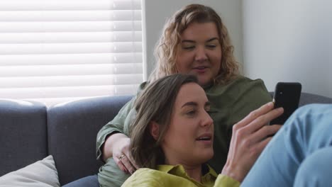 caucasian lesbian couple using smartphone and embracing on couch