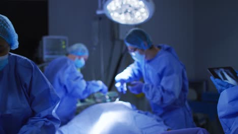 Chirurgen-Mit-Gesichtsmasken-Operieren-Und-Betrachten-Röntgenbilder-Auf-Einem-Tablet-Im-Operationssaal