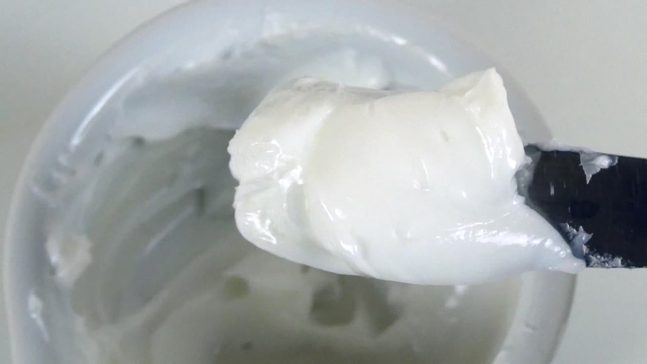 vista macro de manos con guantes en un laboratorio farmacéutico mezclando un ungüento blanco grueso en un mortero de porcelana con un pestillo.