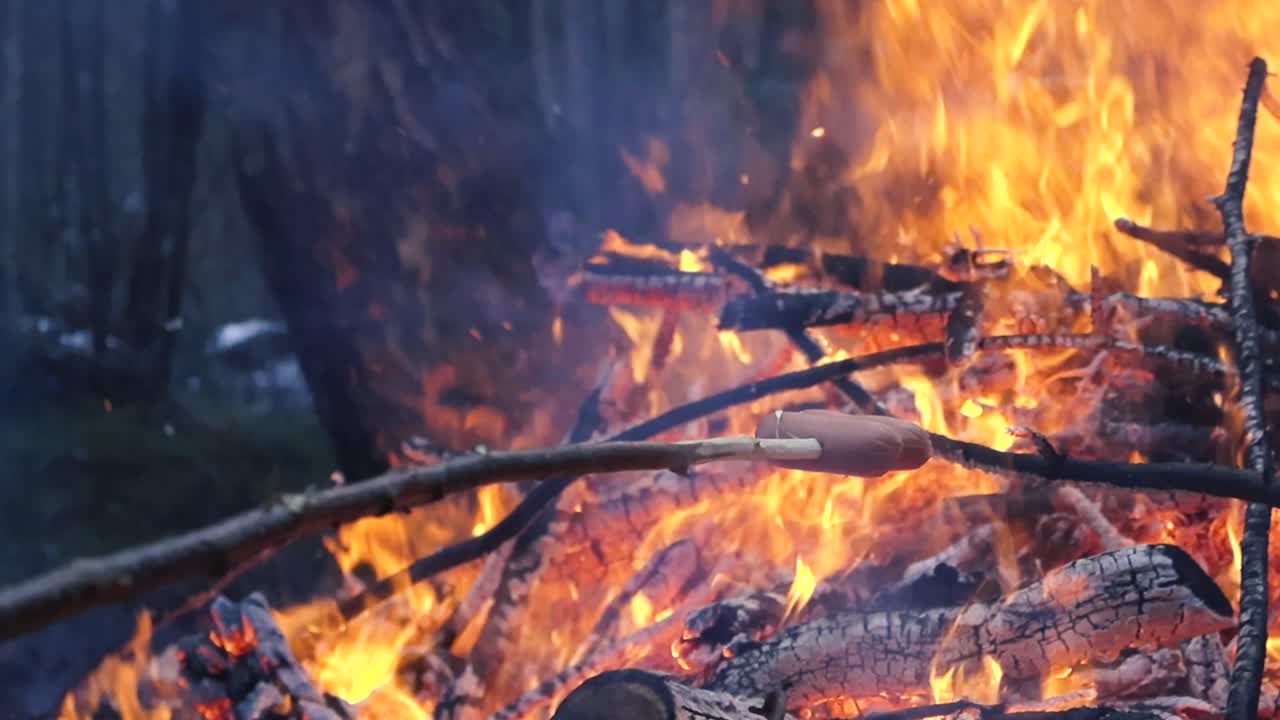 Close up of a fire or a bonfire burning with a large orange vibrant flame in a forest creating thick blue and white smoke while embers fly. Sausage of a hot dog being grilled on fire in slow motion.