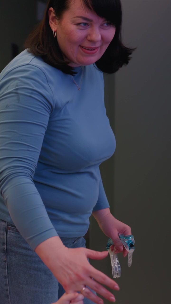Due individui si tengono saldamente in piedi insieme. Una giacca bordeaux e una veste bianca sono appese nelle vicinanze come parte della scena. Pannelli a parete a strisce e vestiti appesi visibili dietro di loro
