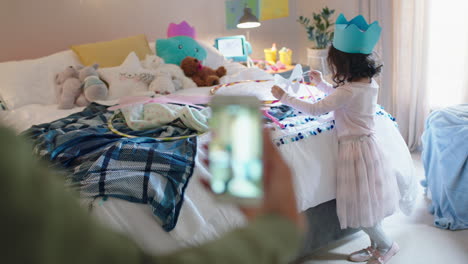 Niña-Feliz-Jugando-A-Vestirse-Con-Máscara-En-El-Dormitorio-Con-Su-Madre-Tomando-Fotos-Usando-Un-Teléfono-Inteligente-Compartido-En-Las-Redes-Sociales