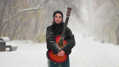 young woman standing in snowy park holding vibrant red acoustic guitar, surrounded by frosty trees and softly falling snow and blur view of people in the distant