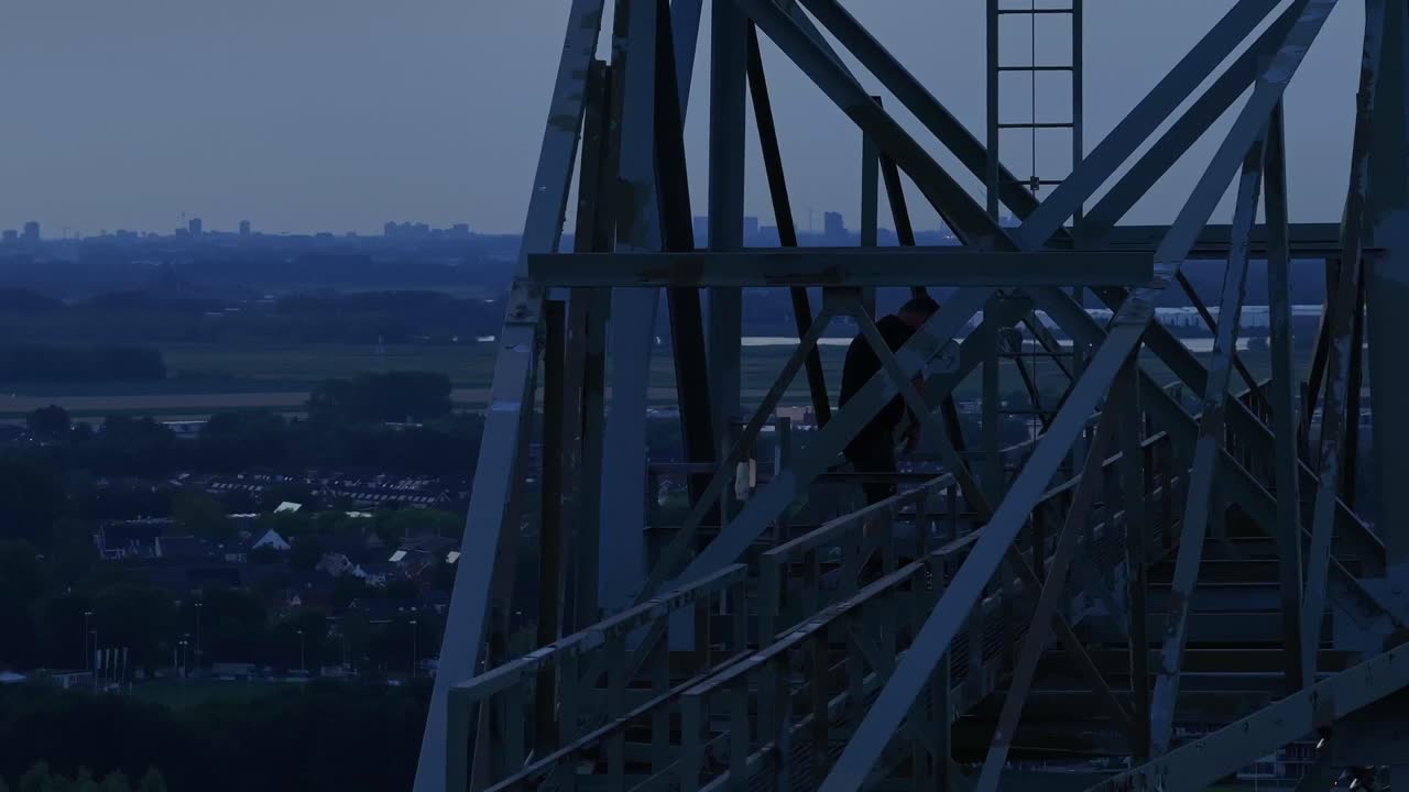 A tall metal structure overlooks a cityscape at dusk, with fields and buildings in the distance
