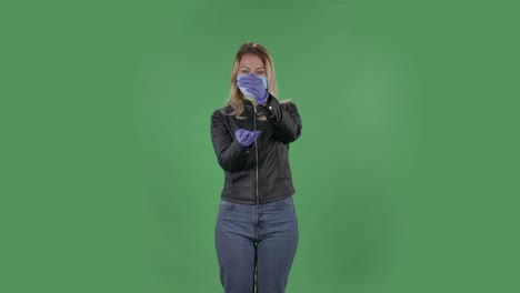 코로나 바이러스의 확산을 막기 위해 의료 보호 얼굴 마스크를 입은 아름다운 젊은 여성의 초상화. 검은 재과 청바지를 입은 느슨한 머리카락을 가진 금발이 스튜디오의 녹색 스크린에 건강 보호 코로나