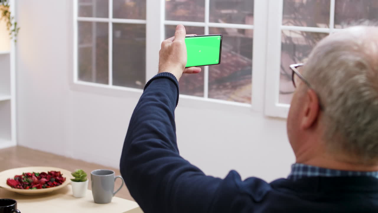 Senior Man Holding Smartphone with Green Screen Indoors