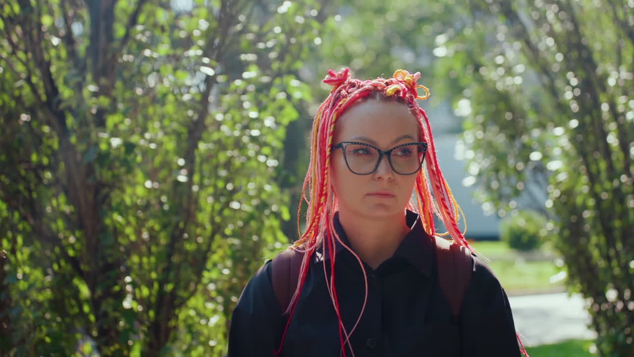 mujer estoica caucásica con trenzas rosas enmarcada por un fondo frondoso, gafas, primer plano con mirada frontal, luz solar suave y bokeh, intensidad silenciosa de una estudiante haciendo una pausa en la naturaleza, escena verde urbana reflexiva