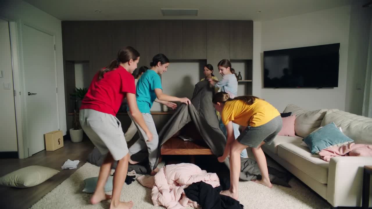 Approaching low table, four teenage females pulling gray blanket from coffee table, creating canopy