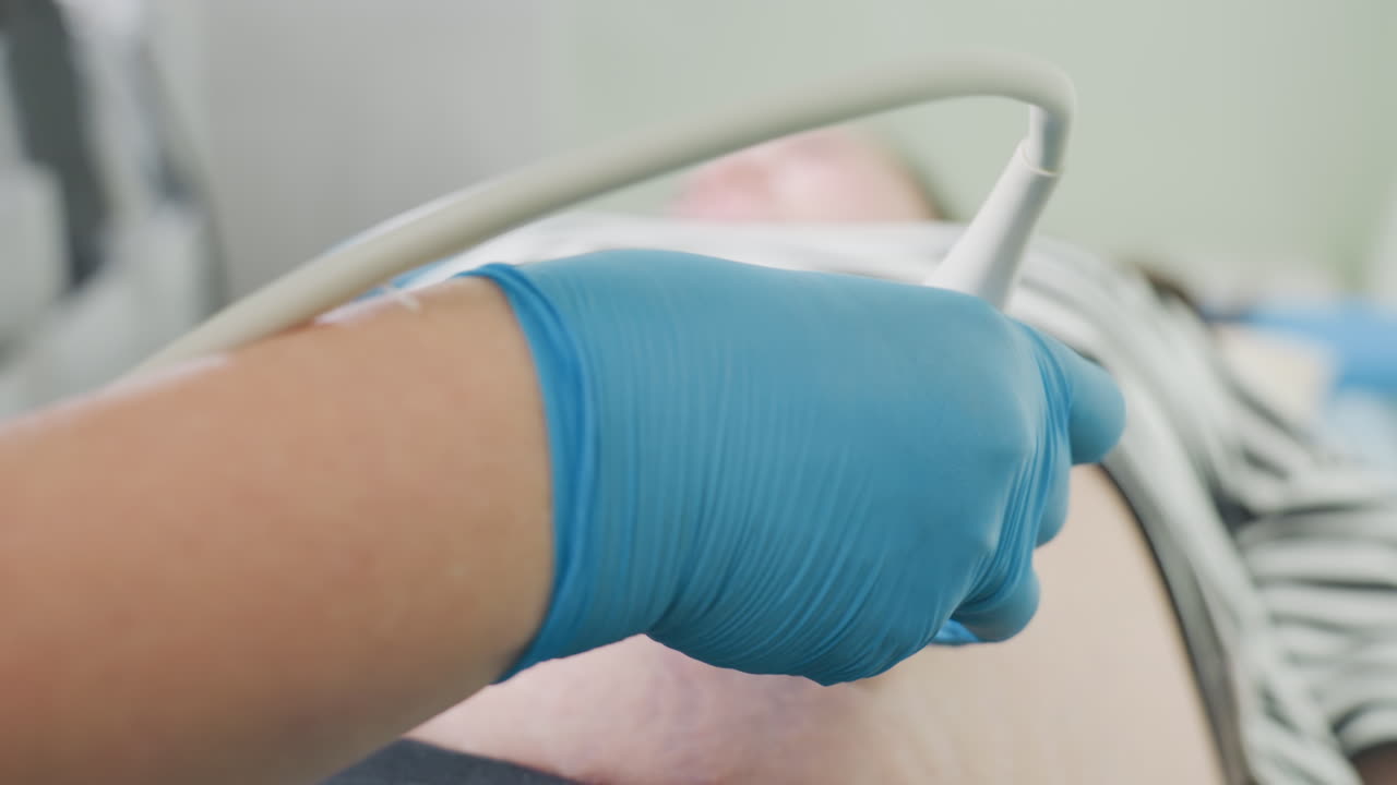 Close up of clinical laboratory scientist wearing sterile gloves moving scanning probe across lower abdomen of patient during diagnostic ultrasound session in clean clinical setting for accurate internal imaging