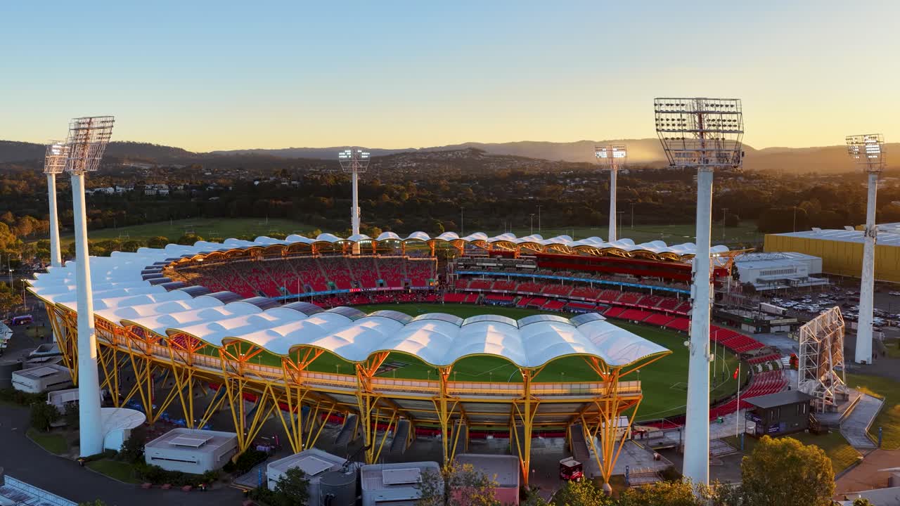 Drone glides over a brightly lit stadium near a river at sunset, revealing architectural details, lush surroundings, and dramatic golden-hour lighting