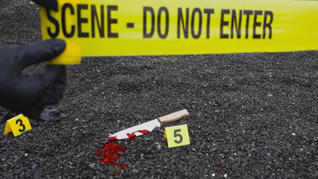 Forensic investigator or police officer with black nylon gloves un rolling and pulling a yellow Crime scene tape in front of a bloody knife and a glove on dark gravel ground next to crime markers, day