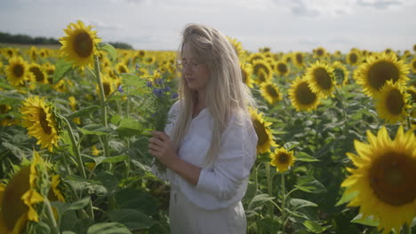 Blonde-with-glasses-in-vivid-yellow-sunflower-field-smells-blue-flowers,-slomo