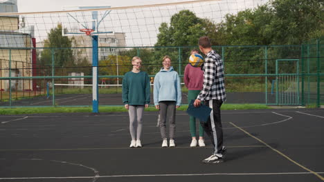 jóvenes jugadores de voleibol se paran en la cancha al aire libre, sonriendo mientras el entrenador rebota el voleibol, la sesión de entrenamiento tiene lugar bajo un cielo despejado, rodeado de árboles, cercas metálicas y edificios residenciales