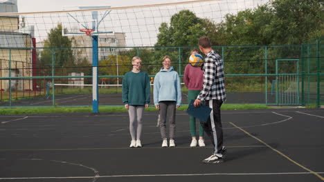 young volleyball players stand on outdoor court, smiling while coach bounces volleyball, training session takes place under clear sky, surrounded by trees, metal fence, and residential buildings