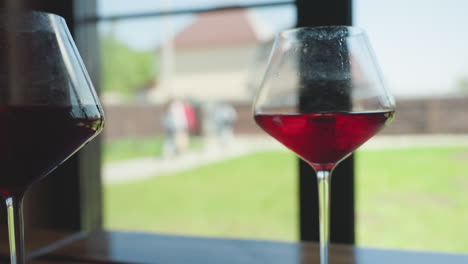 close up of red wine glasses placed on wooden surface by window with soft focus background showing people walking out of compound on sunny day