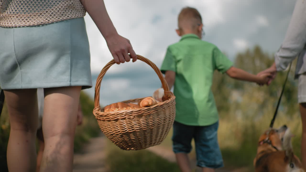 mujer llevando una canasta de bocadillos caminando por un camino de tierra mientras el niño sostiene la mano de su madre, la niña caminando delante, el perro en la correa, rodeado de exuberante vegetación bajo un cielo parcialmente nublado
