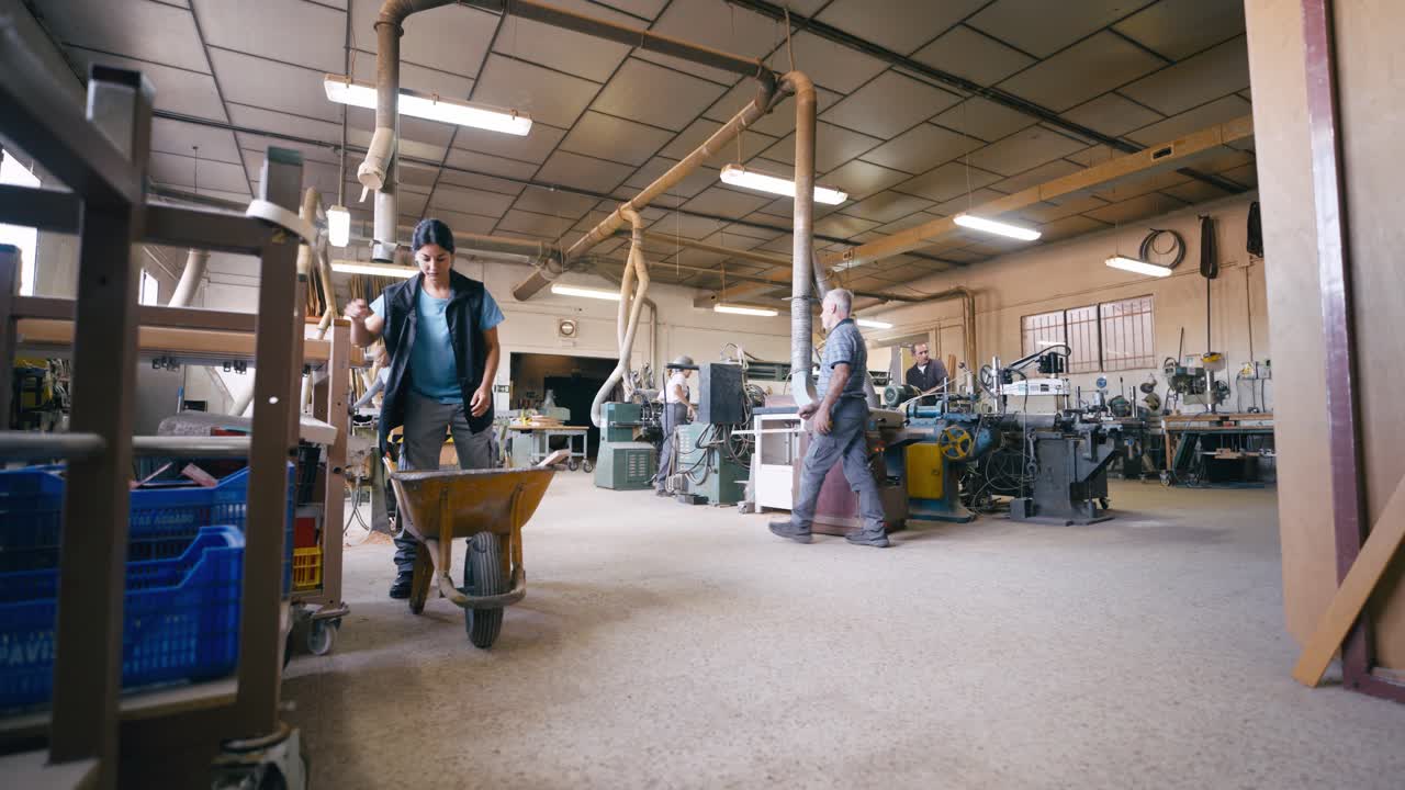 Woman Pushing Wheelbarrow in Busy Woodworking Workshop