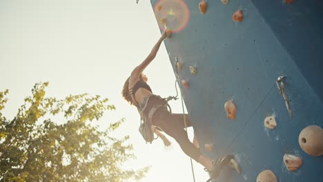 Shot-from-below,-a-blonde-girl-in-a-black-sports-uniform-and-the-necessary-ammunition-and-insurance-climbs-up-the-route-of-a-blue-climbing-wall-on-a-sunny-summer-day