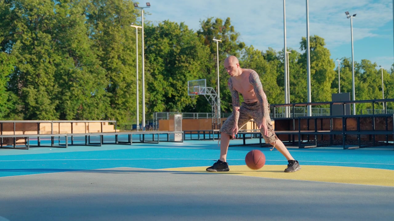 Açık hava sahasında basketbol topu sektiren kel ve dövmeli adam.