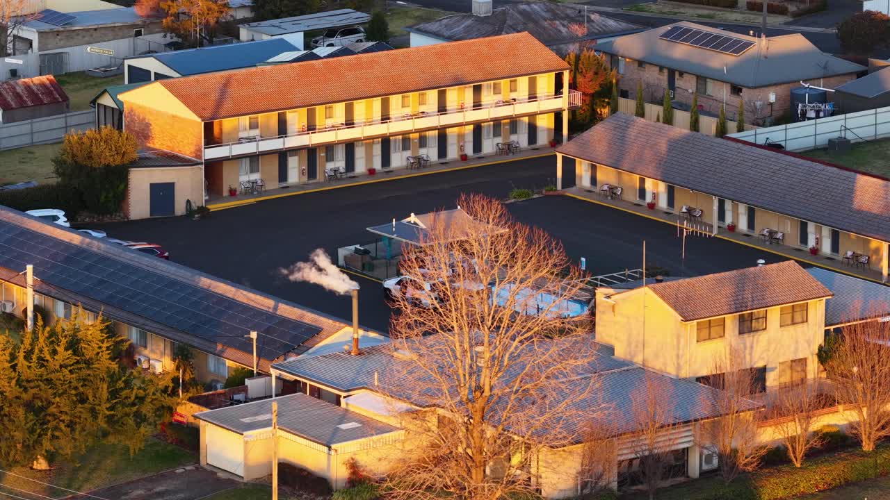 High-angle aerial shot of a quiet town street and buildings at sunset, with smoke drifting from chimneys. Warm golden lighting, minimal camera movement