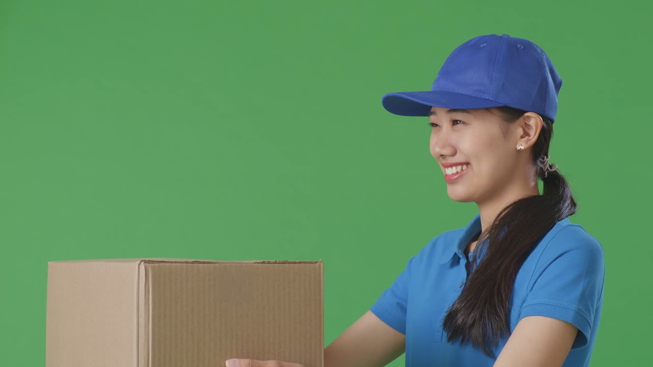 Close Up Of Asian Female Courier In Blue Uniform Giving A Carton To Someone And Smiling While Delivering It On Green Screen Background In The Studio
